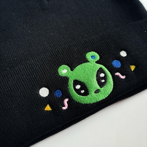 May include: Black beanie featuring an embroidered green alien face with large black eyes and small blue ears. Colorful embroidered accents include white, blue, and yellow shapes. A fun, whimsical accessory.