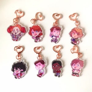 May include: Eight colourful cartoon character keychains with gold heart-shaped clasps. The characters are drawn in a chibi style and include a clown, a girl with red hair, a boy with brown hair, a boy with blonde hair, a boy with black hair, a boy with brown hair, a boy with brown skin, and a boy with blonde hair.