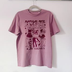 May include: A mauve-colored t-shirt with a graphic featuring three anime-style figures. The text "PUPPYGIRL MOTEL" is above the figures, and "all welcome:3" is below. The shirt is hanging on a gray hanger against a white wall.