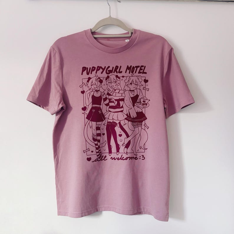 May include: A mauve-colored t-shirt with a graphic featuring three anime-style figures. The text "PUPPYGIRL MOTEL" is above the figures, and "all welcome:3" is below. The shirt is hanging on a gray hanger against a white wall.