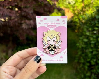 May include: A pink and gold enamel pin featuring a cartoon catboy angel with a halo and wings. The pin has the text "angel catboy enamel pin" and "www.beebeezed.shop" on it.