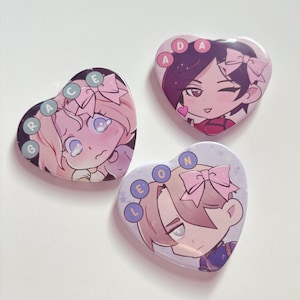 May include: Three heart-shaped pin badges with anime-style character illustrations. The badges feature pink, purple, and blue colour schemes, with the names "Grace", "Ada", and "Leon" encircling the characters. Each character has a bow.