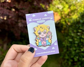 May include: A purple and blue enamel pin featuring a cartoon catboy with yellow hair, wearing a purple and blue outfit and holding a pink heart-shaped lollipop. The pin has the text "magical catboy" and "enamel pin" on it. The text "www.beebeezed.shop" is also on the pin.