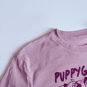 May include: A dusty rose-colored t-shirt with the word "PUPPYG" printed in a dark purple font. The shirt has a crew neck and short sleeves. The graphic is of a cartoon dog.