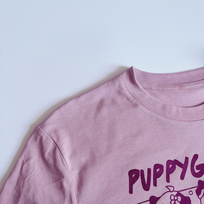 May include: A dusty rose-colored t-shirt with the word "PUPPYG" printed in a dark purple font. The shirt has a crew neck and short sleeves. The graphic is of a cartoon dog.