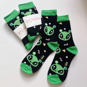 Può includere: Calzini neri con facce di alieni verdi, polsini e dita verdi. I calzini hanno punti bianchi, triangoli gialli e riccioli rosa. Vengono mostrate due paia, con altre due paia avvolte con un'etichetta bianca che dice "socks".