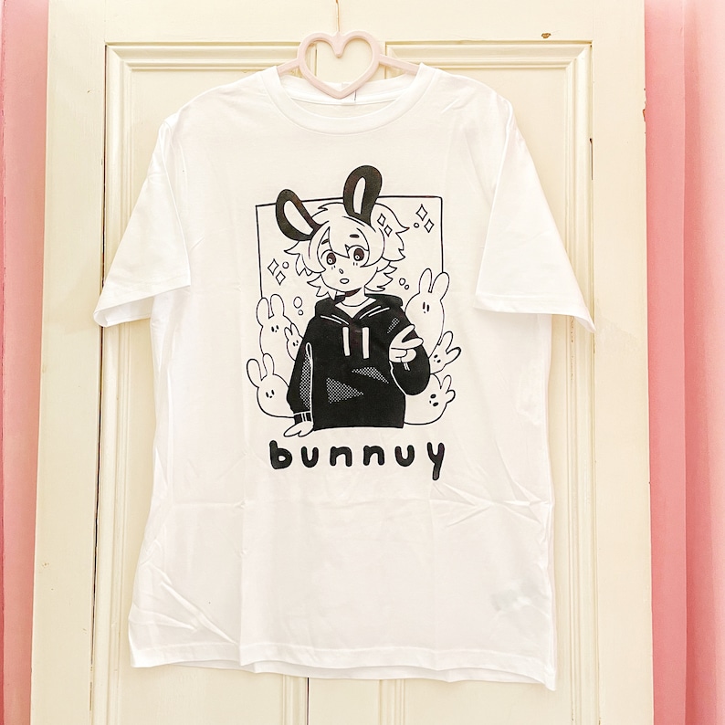 Puede incluir: Camiseta blanca con una ilustraci&oacute;n en blanco y negro de una persona con orejas de conejo que lleva una sudadera con capucha negra y est&aacute; rodeada de conejos. El texto "bunnuy" est&aacute; impreso debajo de la ilustraci&oacute;n.