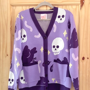 Puede incluir: Un cárdigan lila con un diseño de calaveras y estrellas. El suéter con cuello en V tiene dos bolsillos y una botonadura frontal. Los ribetes y botones son de color púrpura oscuro. La etiqueta dice "bbz shop".
