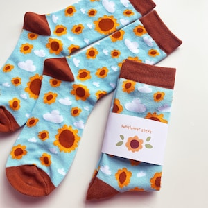 May include: A pair of blue socks with a sunflower and cloud pattern. The socks have brown cuffs and are packaged with a white label that says "Sunflower socks".
