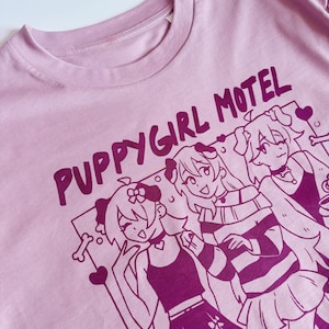 May include: A light pink t-shirt with the text "PUPPYGIRL MOTEL" in dark pink. The shirt features a graphic of three anime-style characters with dog ears and accessories. The design includes hearts and a bone.