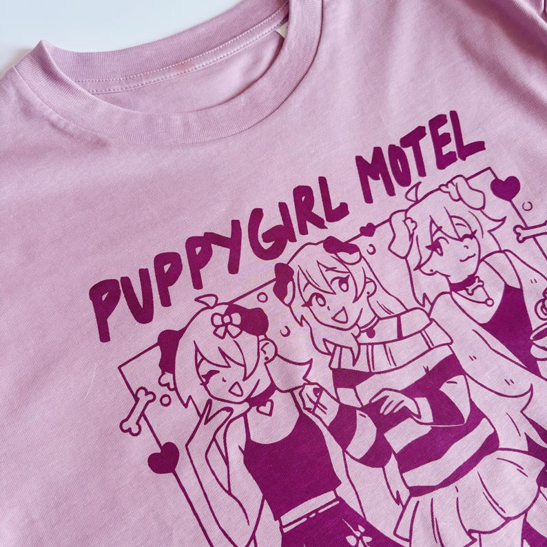 May include: A light pink t-shirt with the text "PUPPYGIRL MOTEL" in dark pink. The shirt features a graphic of three anime-style characters with dog ears and accessories. The design includes hearts and a bone.