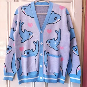 May include: A light purple knit cardigan featuring a repeating pattern of blue sharks and small pink hearts. The garment has a V-neckline, a button-down front with four white buttons, two front pockets, and light blue ribbed trim on the cuffs and hem.