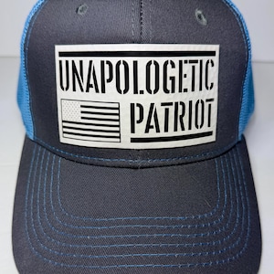 May include: A charcoal grey trucker hat with bright blue mesh back panels and a matching blue stitched brim. A white rectangular patch on the front features the black text UNAPOLOGETIC PATRIOT and a stylized black and white American flag graphic.
