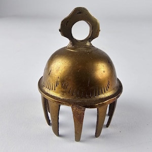 May include: A small, antique brass bell with a rounded top and a decorative loop for hanging. The bell has a series of vertical lines etched around the base and is supported by three curved legs. The bell has a warm, golden hue.