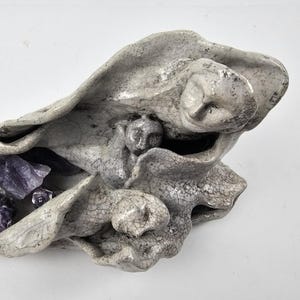 May include: A gray ceramic sculpture with a textured surface, holding several purple amethyst crystals. The sculpture has a unique, organic shape with multiple faces and folds. The crystals are various sizes and shapes, adding a natural contrast to the ceramic.