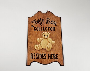 May include: Wooden sign with a brown background and black text. The sign reads "Teddy Bear Collector Resides Here". A painted image of a teddy bear is in the center of the sign.