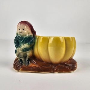 May include: A ceramic planter shaped like a pumpkin with a small gnome figure sitting on the side. The planter is yellow with brown accents and the gnome is wearing a green and blue outfit with a red hat.