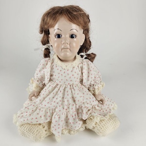 May include: A porcelain doll with brown hair, blue eyes, and a white dress with pink floral print. The doll is wearing crocheted booties.