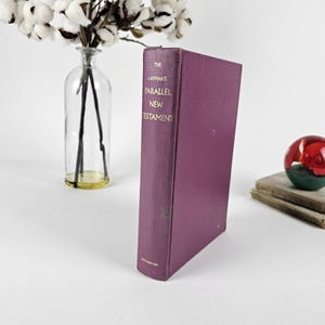 May include: A hardcover book titled "The Layman's Parallel New Testament" in a deep purple color. The book is standing upright, with gold lettering. A glass vase with cotton stems and a decorative glass ball are also in the image.
