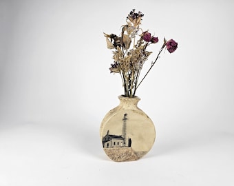 May include: A beige ceramic vase with a lighthouse illustration, holding dried flowers with brown and purple hues. The vase has a round shape and a textured surface, creating a rustic aesthetic.