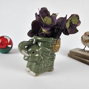 May include: A green ceramic elephant planter holding dark purple flowers. A decorative bird figurine and a red and green glass ball are also present. The planter has a pineapple design on the back.
