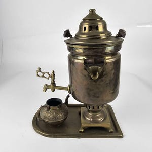 May include: A brass samovar with a small teapot and a tray. The samovar has a round body with a spout and a lid with a handle. The teapot has a round body with a spout and a handle. The tray is rectangular and has a raised edge.