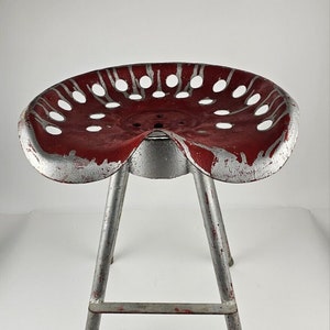 May include: A red and silver metal tractor seat stool with a three-legged base. The seat has a distinctive design with holes and a slight curve in the front.