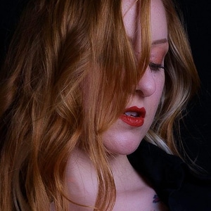 Puede incluir: Una mujer con el pelo largo, ondulado y castaño rojizo mira hacia abajo. Su cabello cubre la mayor parte de su rostro. Lleva lápiz labial rojo y una camisa negra.
