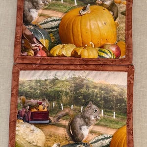 May include: Two square fabric pot holders with a brown quilted border. The top features a large orange pumpkin and two squirrels. The bottom shows a red truck filled with pumpkins, a squirrel, and various gourds in a field setting.