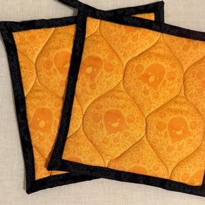 May include: Two square quilted pot holders featuring an orange fabric with a repeating pattern of small smiling ghosts and circular shapes. The pot holders are finished with a solid black fabric binding along all four edges and a hanging loop.