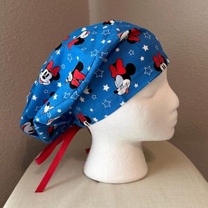 May include: A blue scrub cap featuring a pattern of Minnie Mouse, red bows, and white stars. The cap has red ties and is displayed on a white mannequin head. The fabric is a bright blue.