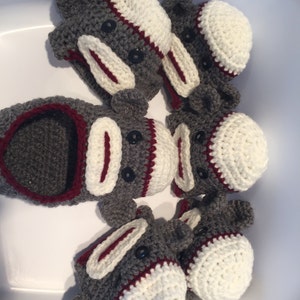 May include: A collection of handmade sock monkey toys. Each monkey is crocheted with gray yarn, featuring white and burgundy accents on the face and hat. Black button eyes and a red mouth complete the design. The toys are arranged in a circular pattern.