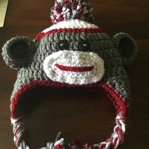 May include: A gray, red, and white crocheted monkey hat with a pom-pom on top. The hat has a long, braided tie.