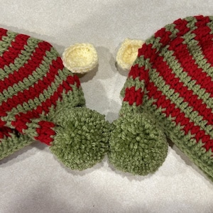 May include: Two handmade crochet elf hats in red and green stripes, with green pom-poms and small cream-colored circles. The hats are designed with a long, pointed shape, perfect for festive occasions.