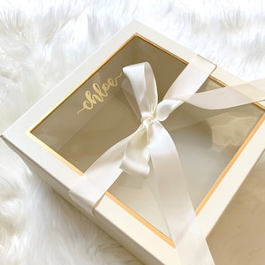 May include: A white gift box with a clear window and a gold trim. The box is tied with a white satin ribbon and the name "Chloe" is written in gold lettering on the top of the box.
