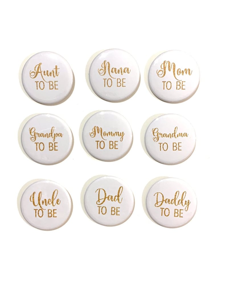 May include: Nine white round buttons with gold lettering. The buttons say "Aunt to Be", "Nana to Be", "Mom to Be", "Grandpa to Be", "Mommy to Be", "Grandma to Be", "Uncle to Be", "Dad to Be", and "Daddy to Be".