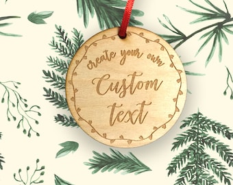 May include: A round wooden ornament with the words "create your own Custom text" engraved on it. The ornament has a decorative border and a red ribbon for hanging. The background features a pattern of green watercolor pine branches and leaves.