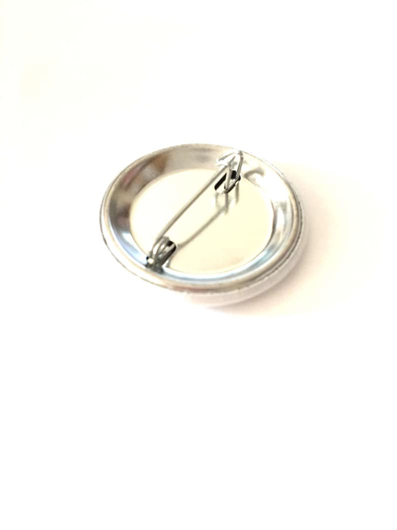 May include: The back of a silver button with a safety pin attached. The button is blank and ready to be customized.