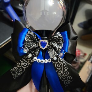 May include: A black and blue light stick with a large bow. The bow features black ribbon with a silver swirl design and blue ribbon accents. A heart-shaped jewel and the name "JIMIN" are also visible.