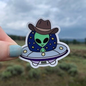 May include: A green alien wearing a brown cowboy hat is flying in a white and purple spaceship with a blue and white starry background. The spaceship has the word "Howdy" written on the bottom.