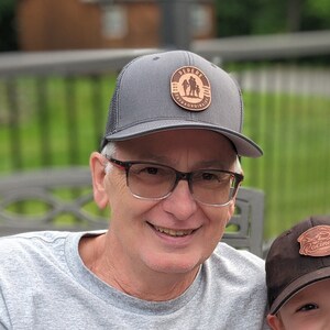 Puede incluir: Un hombre y un niño pequeño con gorras de béisbol grises y marrones con parches de cuero. El hombre lleva una camiseta gris con la palabra "Cornwall" impresa. El niño lleva una camiseta gris con un gráfico de un camión rojo.