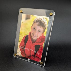 Peut inclure: Un cadre photo en acrylique transparent avec un accent en métal doré sur chaque coin. Le cadre contient une photo d'un jeune garçon portant un sweat-shirt rouge et souriant. La photo porte le texte "Je t'aime Père".