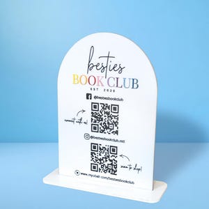 May include: White sign with a rounded top, featuring the text "besties BOOK CLUB EST 2025" in rainbow colours. The sign also includes two QR codes, one for Facebook and one for Instagram, with instructions to "connect with us" and "next to adopt". The sign is on a white stand.