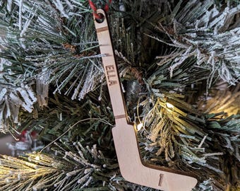 May include: A wooden hockey stick ornament with the name "Ella" engraved on it. The ornament is hanging on a Christmas tree.