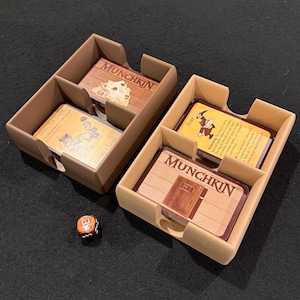 May include: Two light brown plastic card organizers containing stacks of Munchkin game cards. One organizer holds a card labeled Munchkin and a Steal The Credit card. The other holds a Munchkin card and a Thief card. A single orange six-sided die is nearby.