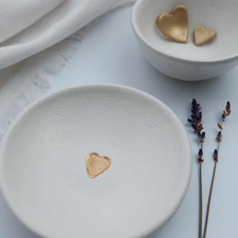 May include: Two white ceramic dishes with gold heart accents. The dish on the left has one gold heart in the center. The dish on the right has two gold hearts.
