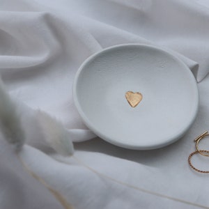 May include: White ceramic ring dish with a gold heart design. The dish is on a white fabric background.