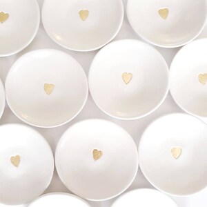 May include: A set of ten white ceramic ring dishes with a gold heart design. The dishes are arranged in a circular pattern on a white background.