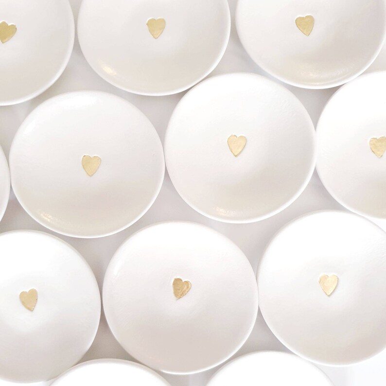 May include: A set of ten white ceramic ring dishes with a gold heart design. The dishes are arranged in a circular pattern on a white background.