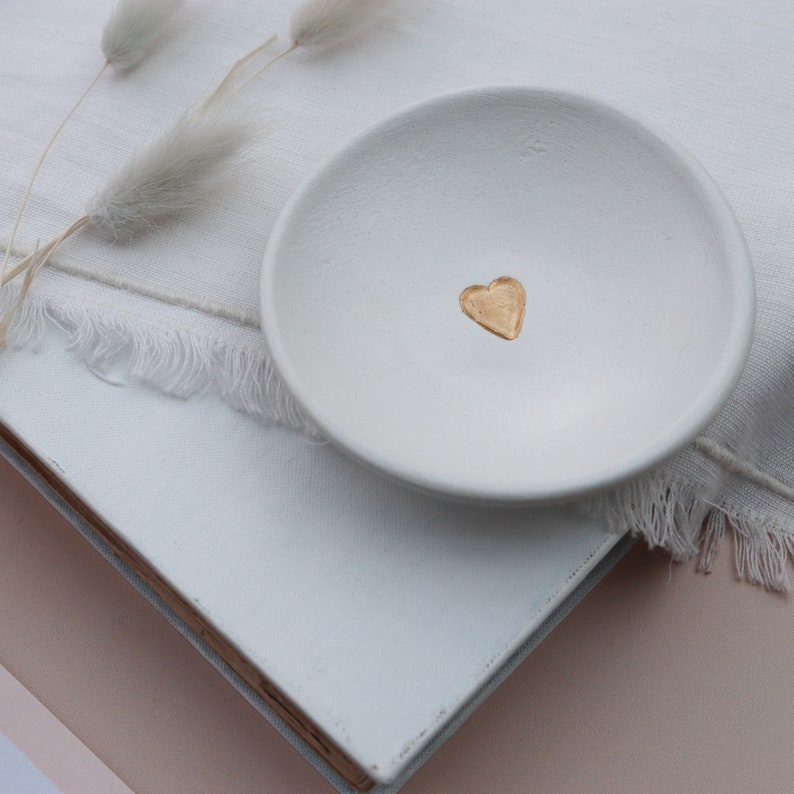 May include: A white ceramic trinket dish with a gold heart design. The dish is sitting on a white surface.